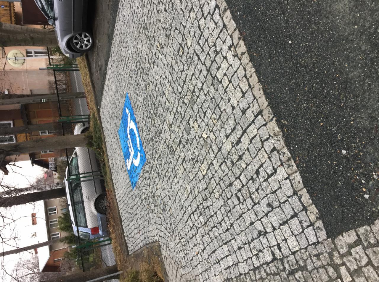 Miejsce parkingowe dla niepełnosprawnych wyłożone kostką brukową w odcieniach szarości, z wyraźnie odmalowanym niebieskim symbolem. Widoczne zaparkowane samochody i otoczenie budynku.