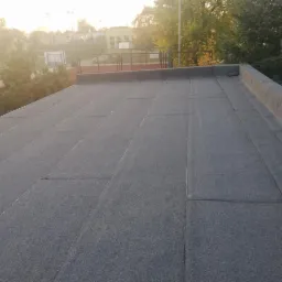 Płaski dach pokryty papą termozgrzewalną, widoczne łączenia arkuszy, w tle zieleń drzew i budynek z kortem tenisowym.