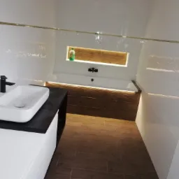 Nowoczesna łazienka z białą armaturą, drewnopodobnymi płytkami i oświetleniem LED, widoczny płyn do mycia w niszy nad wanną.