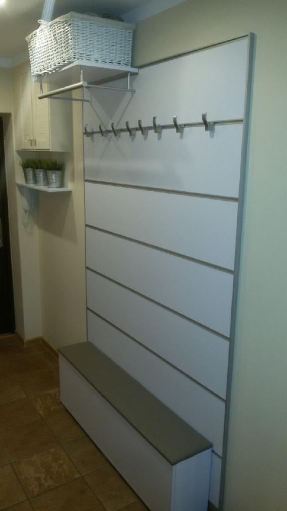 Biała garderoba z wieszakami, półką i siedziskiem w przedpokoju, na górze kosz z wikliny, a na półce po lewej trzy doniczki z zielonymi roślinami.