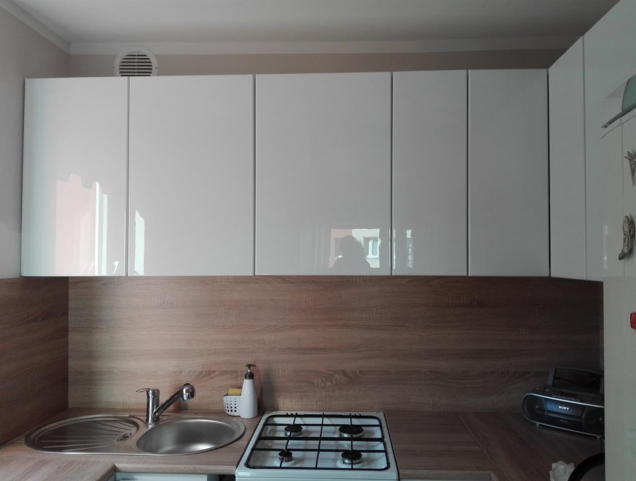 Białe, lakierowane szafki kuchenne wiszące nad blatem z drewnopodobnym dekorem, widoczny zlew, bateria i płyta gazowa.