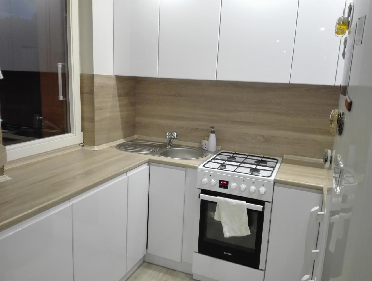 Nowoczesna, jasna kuchnia z białymi, błyszczącymi szafkami i blatem imitującym drewno, widoczna zlew, kuchenka gazowa z ręcznikiem na drzwiczkach piekarnika i okno z widokiem na zewnątrz.