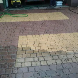 Częściowo umyta kostka brukowa w kolorze brązowo-żółtym, widoczne różnice w czystości, przewód ciśnieniowy i fragment myjki ciśnieniowej w tle.