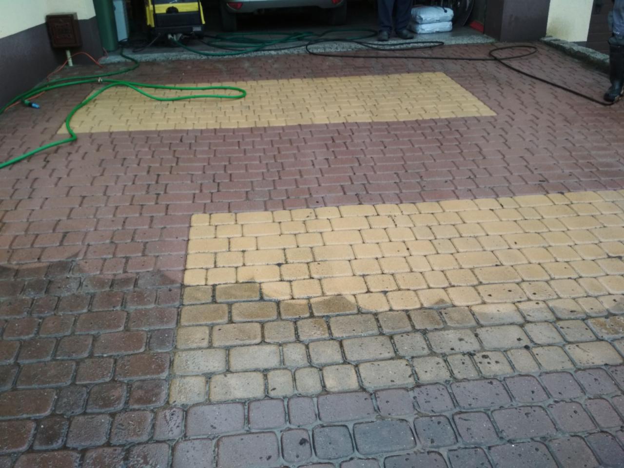 Częściowo umyta kostka brukowa w kolorze brązowo-żółtym, widoczne różnice w czystości, przewód ciśnieniowy i fragment myjki ciśnieniowej w tle.