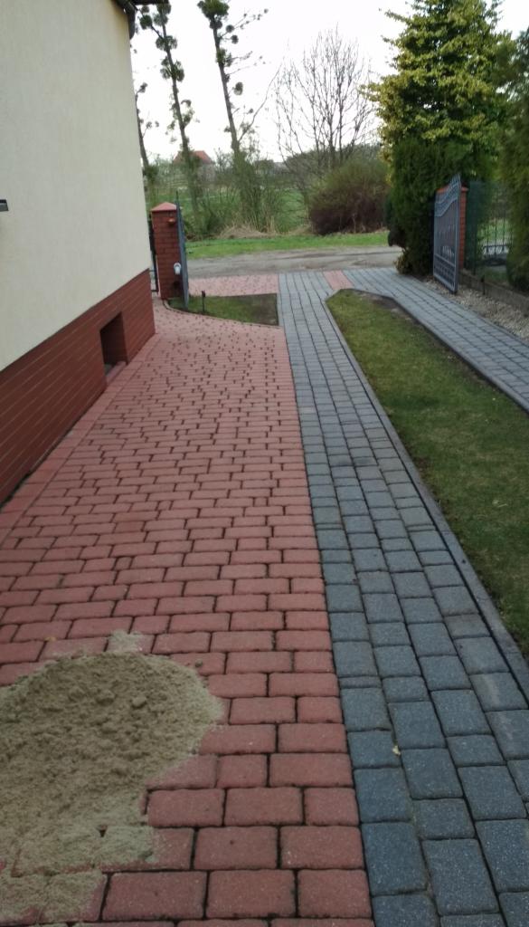 Ułożona kostka brukowa w dwóch kolorach (czerwonym i szarym) prowadząca do bramy, z widocznym piaskiem na czerwonej części, sugerującym prace wykończeniowe.