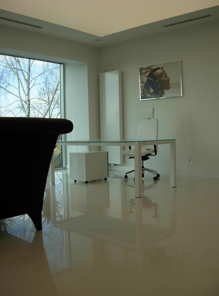 Minimalistyczne biuro z białym biurkiem ze szklanym blatem, krzesłem na kółkach i szafką pod biurkiem, oświetlone dużym oknem z widokiem na drzewo bez liści.