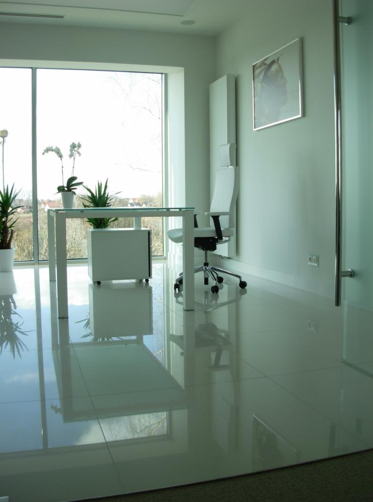 Minimalistyczne biuro z białym biurkiem, szklanym blatem i krzesłem obrotowym na tle dużego okna z widokiem na zewnątrz, uzupełnione roślinami doniczkowymi i abstrakcyjnym obrazem na ścianie.