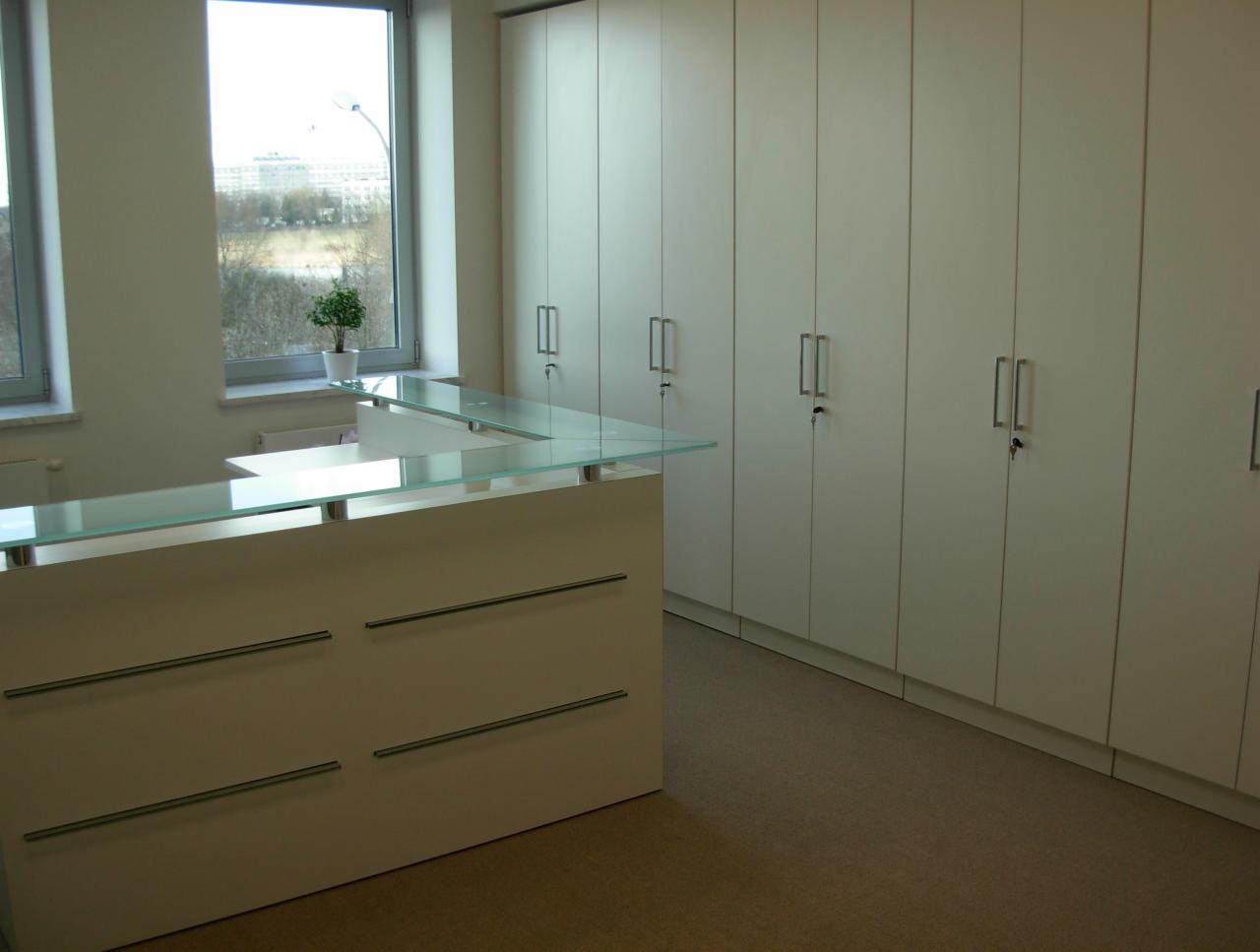 Nowoczesne biuro w Śremie: biała lada recepcyjna z przezroczystym blatem, rząd szafek zamykanych na klucz i widok na zieleń za oknem.