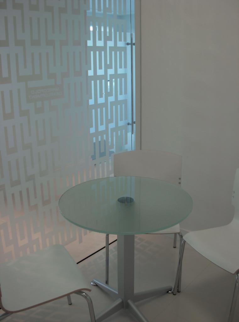 Szklany okrągły stół z metalową podstawą i białymi krzesłami w minimalistycznym biurze z dekoracyjną szybą z geometrycznym wzorem.