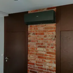 Czarna jednostka klimatyzacyjna Haier zamontowana na ścianie z czerwonej cegły w nowoczesnym wnętrzu z ciemnobrązowymi panelami i białymi ścianami.