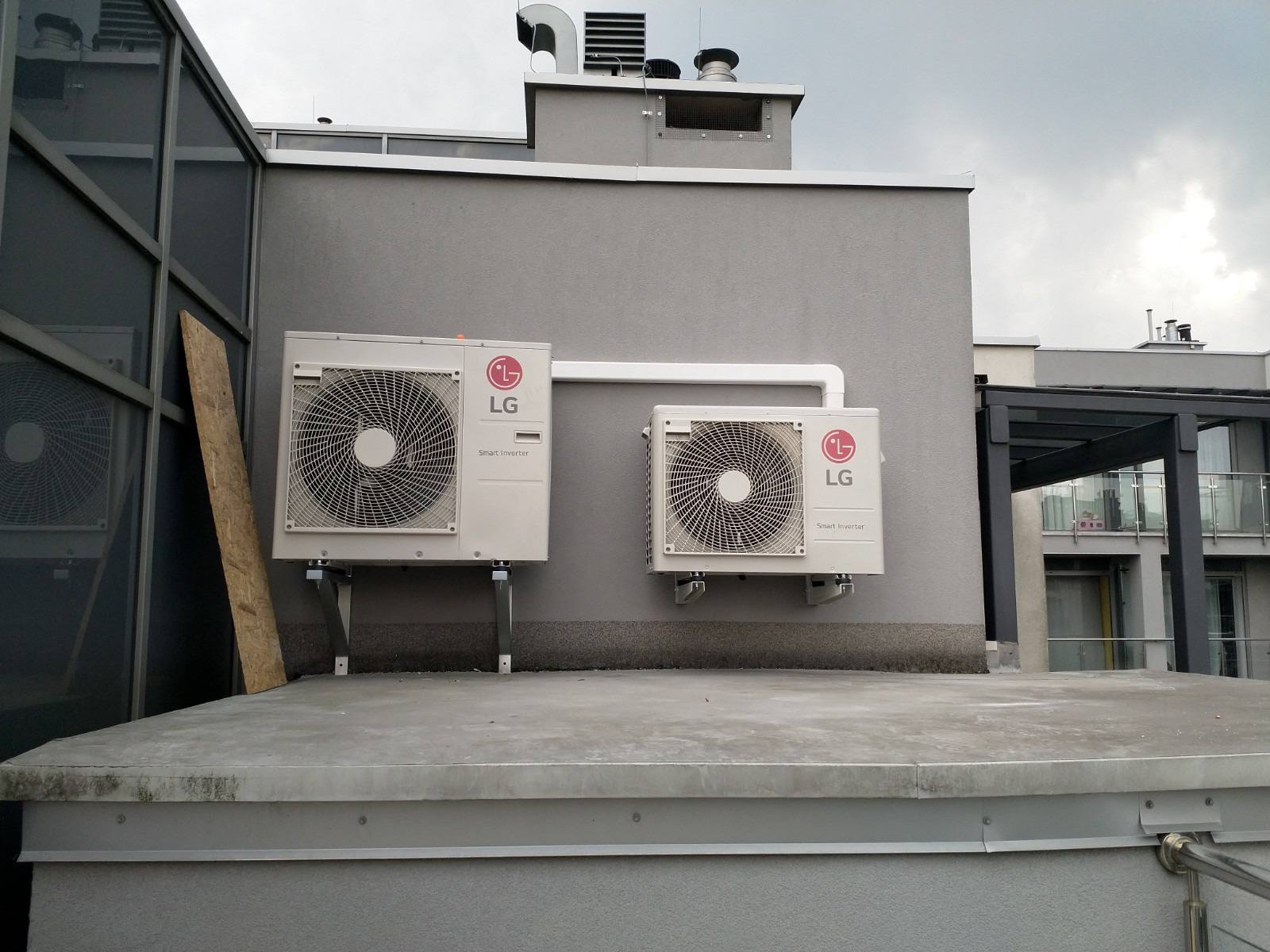 Dwa zewnętrzne moduły klimatyzacji LG Smart Inverter zamontowane na szarym budynku, połączone białymi rurami, widok z dachu, pochmurne niebo w tle.