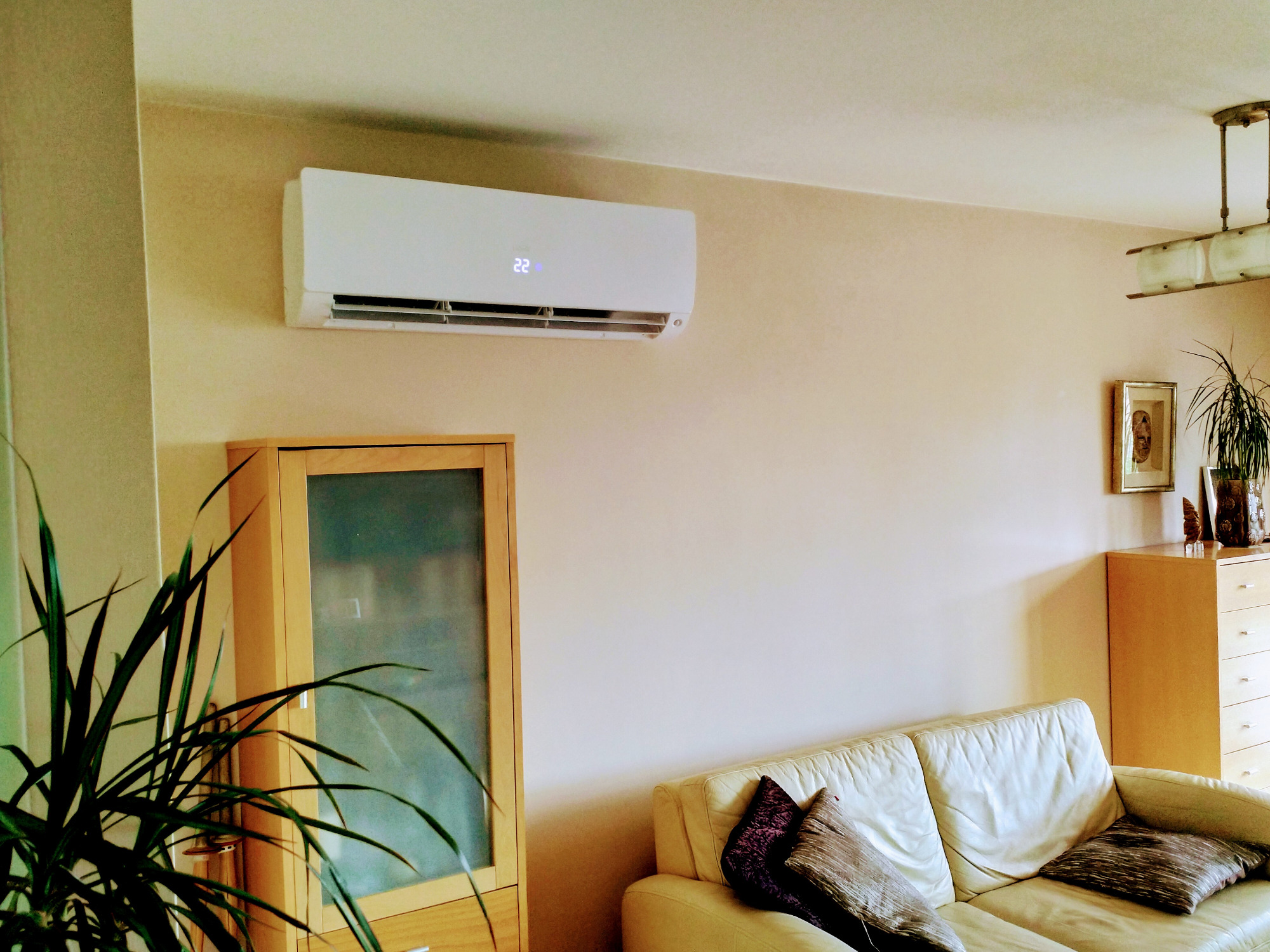 Nowoczesny, biały klimatyzator typu split zamontowany na ścianie w jasnym salonie z beżowymi ścianami, widoczny narożnik sofy i meble.