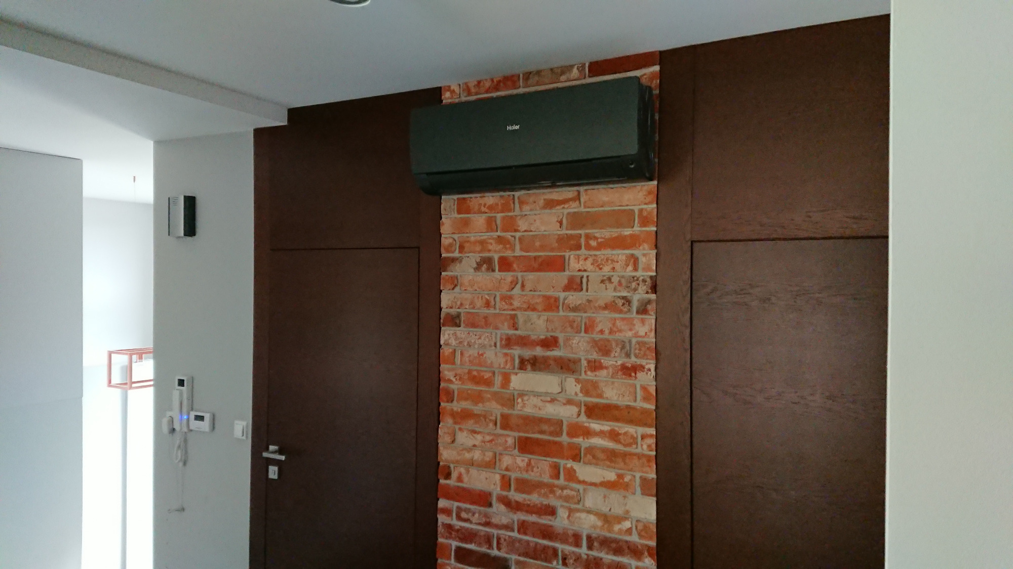 Czarna jednostka klimatyzacyjna Haier zamontowana na ścianie z czerwonej cegły w nowoczesnym wnętrzu z ciemnobrązowymi panelami i białymi ścianami.