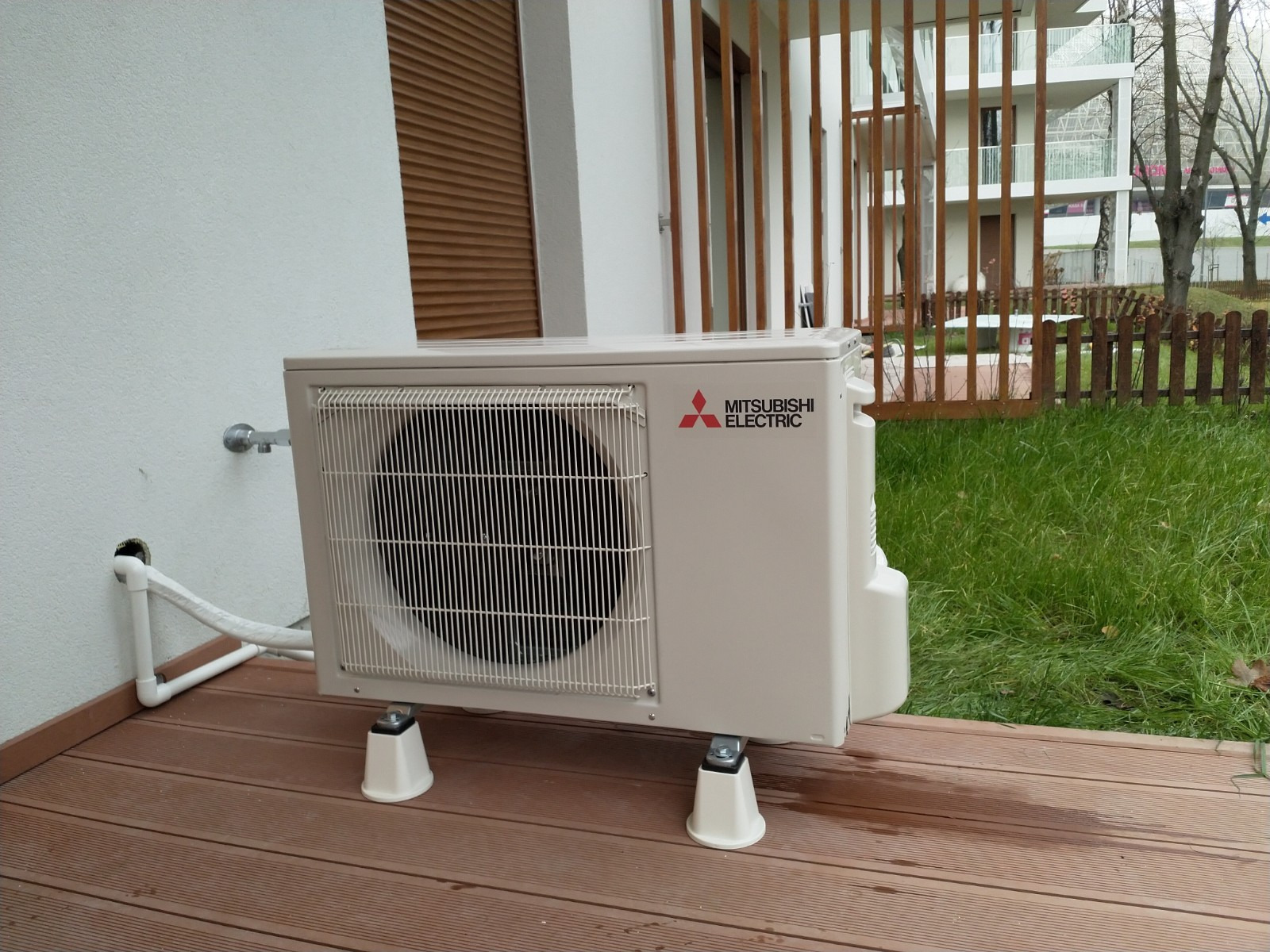 Jednostka zewnętrzna klimatyzatora Mitsubishi Electric na drewnianym tarasie, z widocznymi rurami odprowadzającymi skropliny i elementami montażowymi, na tle budynku z drewnianymi żaluzjami...