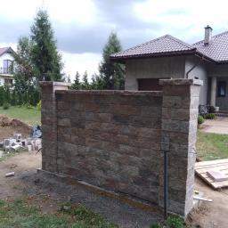 Grzegorz Wróbel - Budowa ogrodzenia z bloczków betonowych w trakcie realizacji, widoczny fragment muru, niezabezpieczone przewody elektryczne i ułożone bloczki w tle, dom jednorodzinny w tle.