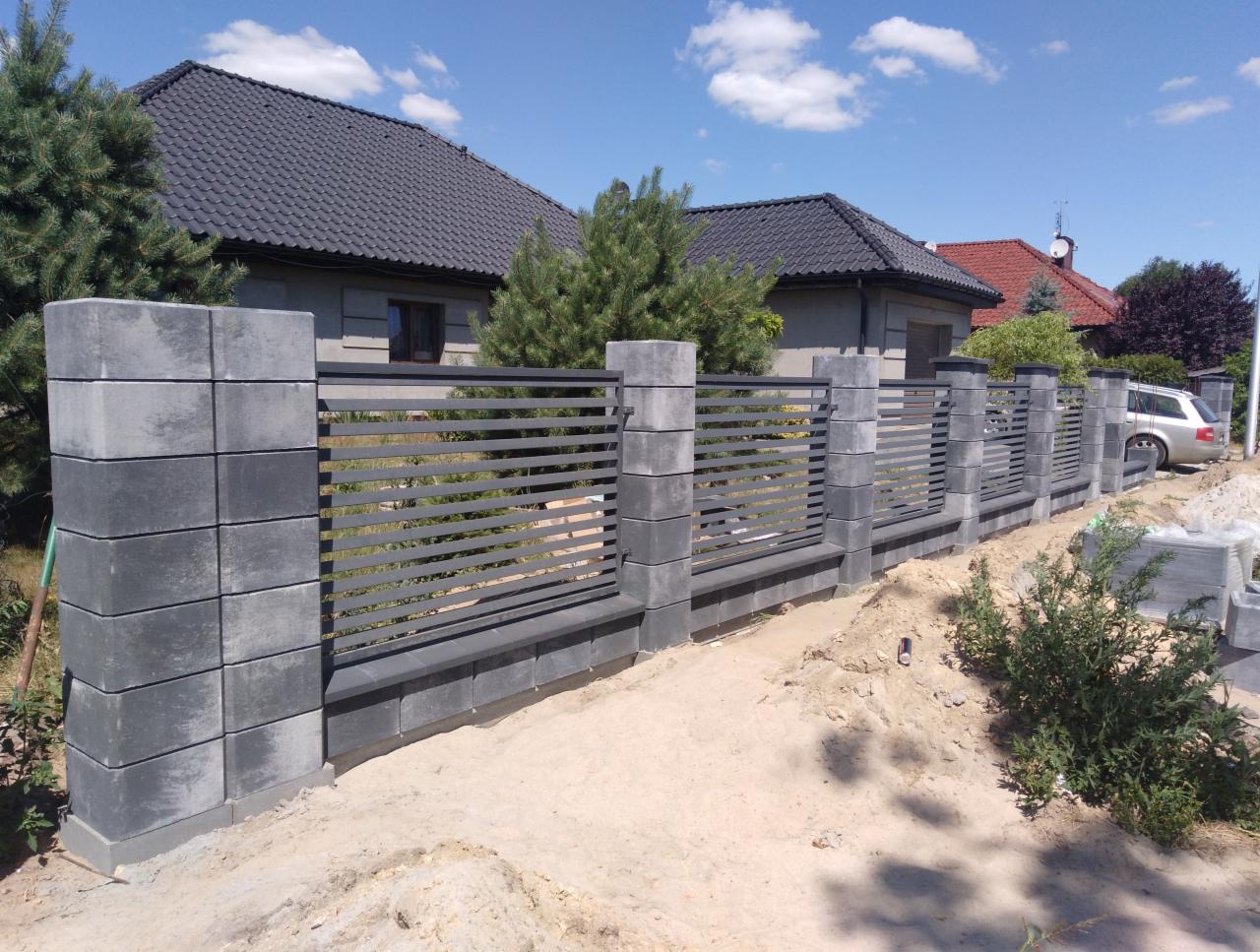Nowoczesne ogrodzenie posesji z szarych betonowych słupków i poziomych metalowych przęseł, widok z zewnątrz na tle domów jednorodzinnych i błękitnego nieba.