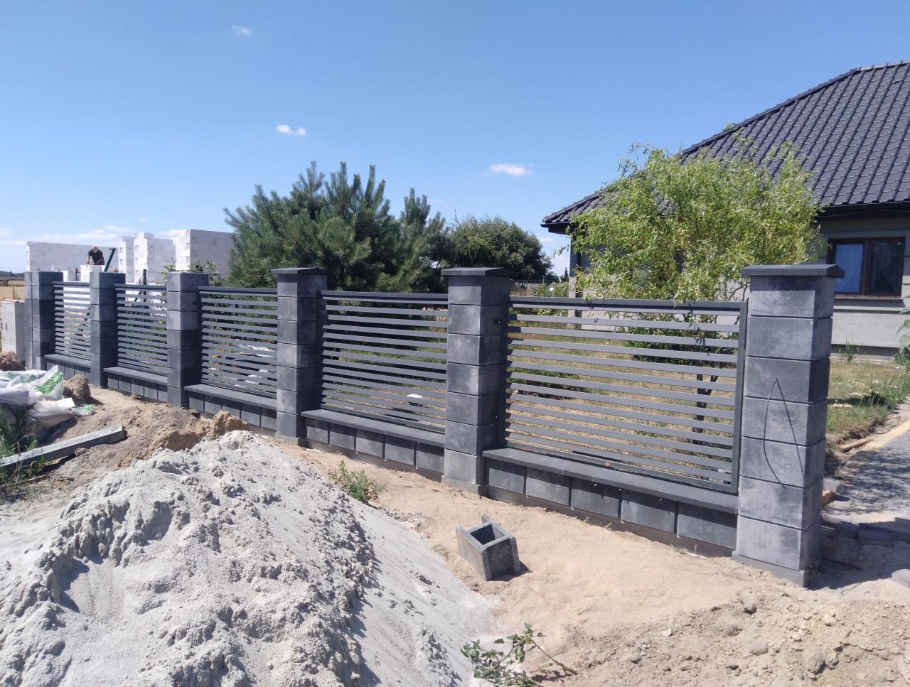 Nowoczesne ogrodzenie panelowe z elementami betonowymi, widoczne ślady prac ziemnych i budowlanych, dom w tle.