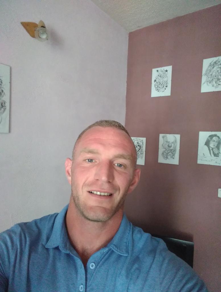 Uśmiechnięty mężczyzna w niebieskiej koszuli na tle ściany w dwóch odcieniach z rysunkami i lampą