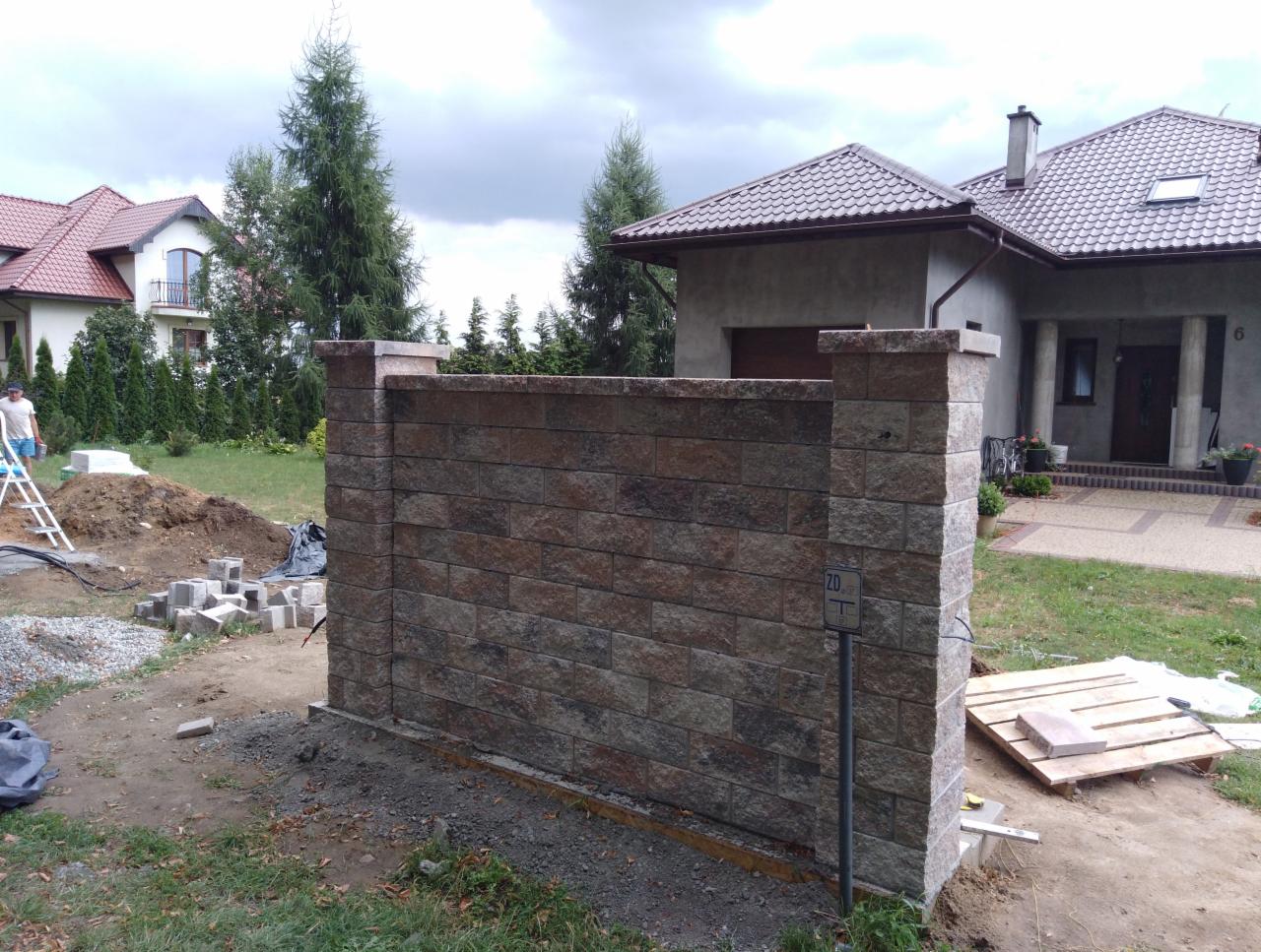 Budowa ogrodzenia z bloczków betonowych w trakcie realizacji, widoczny fragment muru, niezabezpieczone przewody elektryczne i ułożone bloczki w tle, dom jednorodzinny w tle.