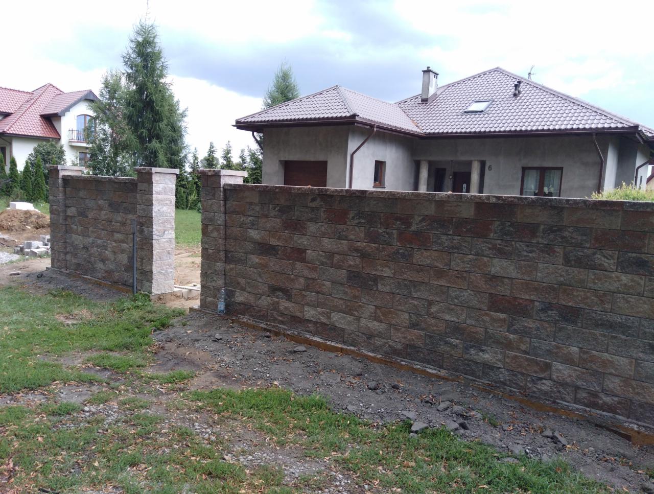 Nowoczesne ogrodzenie z brązowej cegły klinkierowej z jasnymi filarami na tle domu jednorodzinnego z brązowym dachem, widoczne elementy montażowe w ziemi.