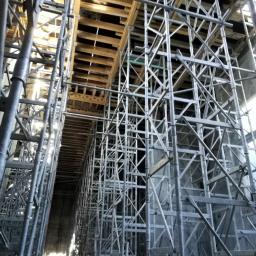 Anna Szczerbal Budownictwo - Skomplikowany system rusztowań podtrzymujących drewniane szalunki stropu na placu budowy, widok z dołu ukazujący perspektywę i konstrukcję wsporczą.