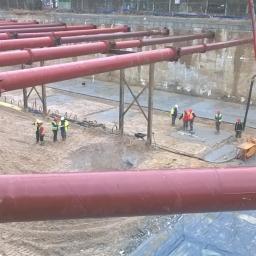 Anna Szczerbal Budownictwo - Montaż rurociągu na placu budowy, widoczni pracownicy w kaskach i kamizelkach odblaskowych, betonowa podstawa i rury w kolorze czerwonym, w tle ściana wykopu.