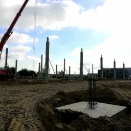 Budowa hali: betonowe fundamenty z wystającymi prętami zbrojeniowymi, w tle stalowe słupy konstrukcyjne i czerwony dźwig na placu budowy.