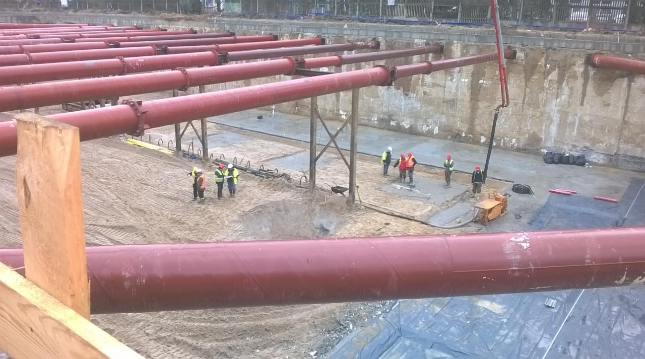 Montaż rurociągu na placu budowy, widoczni pracownicy w kaskach i kamizelkach odblaskowych, betonowa podstawa i rury w kolorze czerwonym, w tle ściana wykopu.