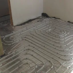 Białe rury ogrzewania podłogowego ułożone w spiralny wzór na siatce, widoczne przygotowania do wylewki, surowe wykończenie ścian.