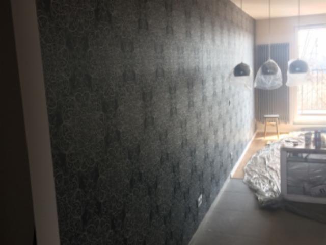 Ściana pokryta ciemną tapetą we wzory, z trzema lampami wiszącymi w tle, zabezpieczonymi folią malarską, widoczny grzejnik i okno w oddali, podłoga zabezpieczona folią.