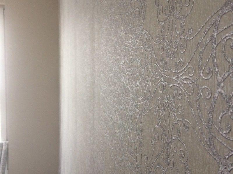Fragment ściany pokrytej tapetą w odcieniach szarości z delikatnym, ornamentacyjnym wzorem w stylu retro, widoczne przejście do gładkiej, jasnej ściany w rogu pomieszczenia.