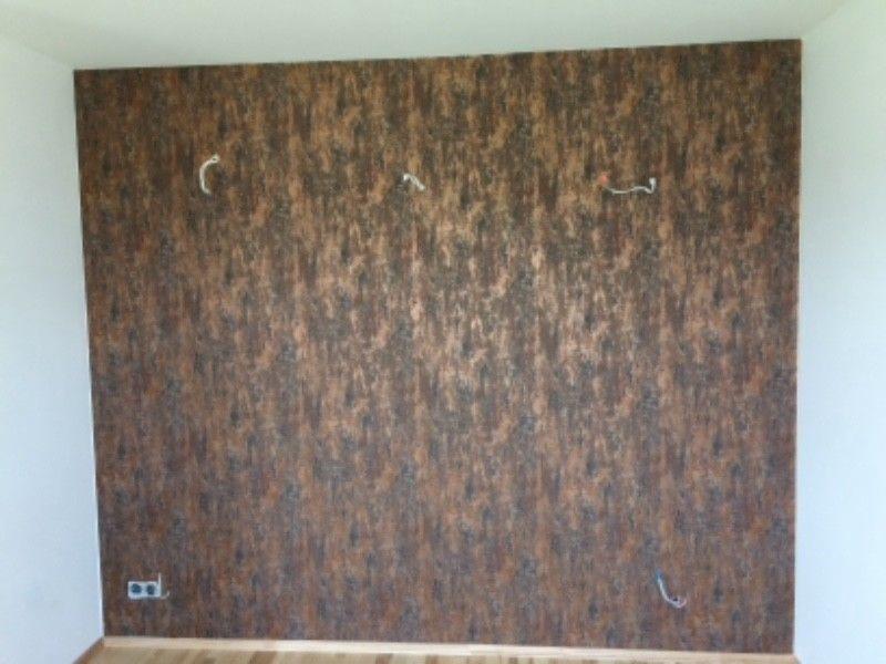 Ściana pokryta tapetą o wzorze imitującym korę drzewa, z wystającymi przewodami elektrycznymi, widoczny fragment podłogi i białe ściany boczne.