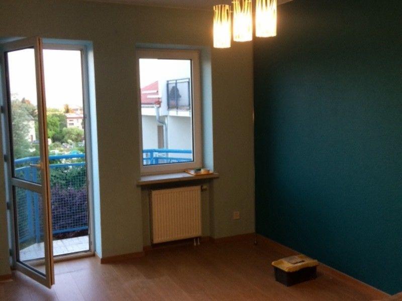 Pomieszczenie po malowaniu: ściana w odcieniu ciemnej zieleni, drewniana podłoga, okno z widokiem na balkon i budynek, grzejnik pod oknem, lampa sufitowa z trzema kloszami, drabinka malarska w rogu...