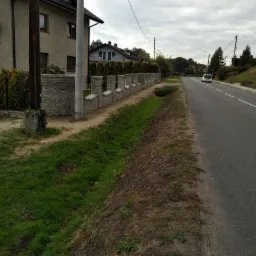 Długi, kamienny mur z dekoracyjnymi elementami, oddzielający posesję od drogi, z widocznym samochodem w oddali.