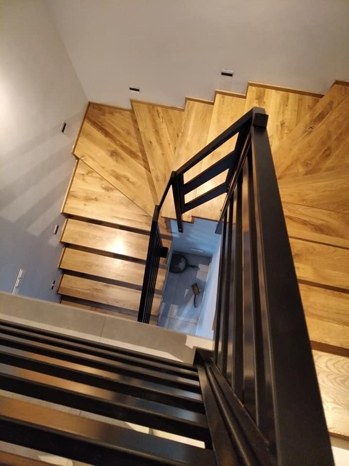 Widok z góry na drewniane schody spiralne z czarną, metalową balustradą, widoczne narzędzia malarskie na podeście.