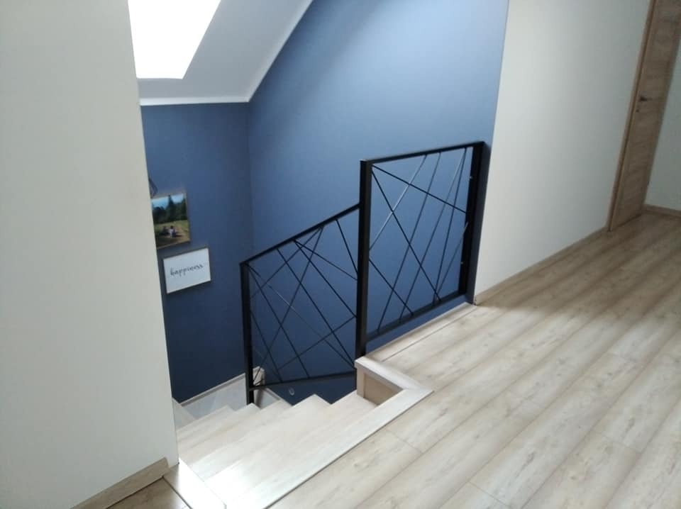 Nowoczesna, czarna poręcz schodowa z geometrycznym wzorem na tle niebieskiej ściany i jasnych drewnianych schodów.