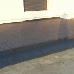 Nowo ułożony, dwupoziomowy taras z ciemnoszarej kostki brukowej przed budynkiem w trakcie wykańczania, widoczne deski i piasek.