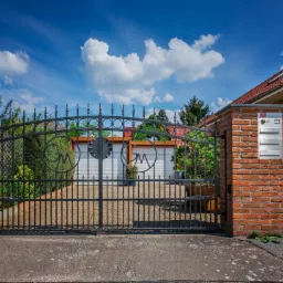 Kuta, czarna brama wjazdowa z dekoracyjnymi elementami w kształcie liter w okręgach, zamontowana między murowanymi z czerwonej cegły słupkami, na tle domu z czerwoną dachówką i błękitnego nieba...