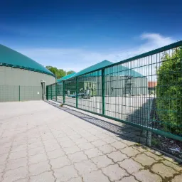 Otwarta metalowa brama panelowa w kolorze zielonym, prowadząca na teren biogazowni z widocznymi zbiornikami i instalacjami.