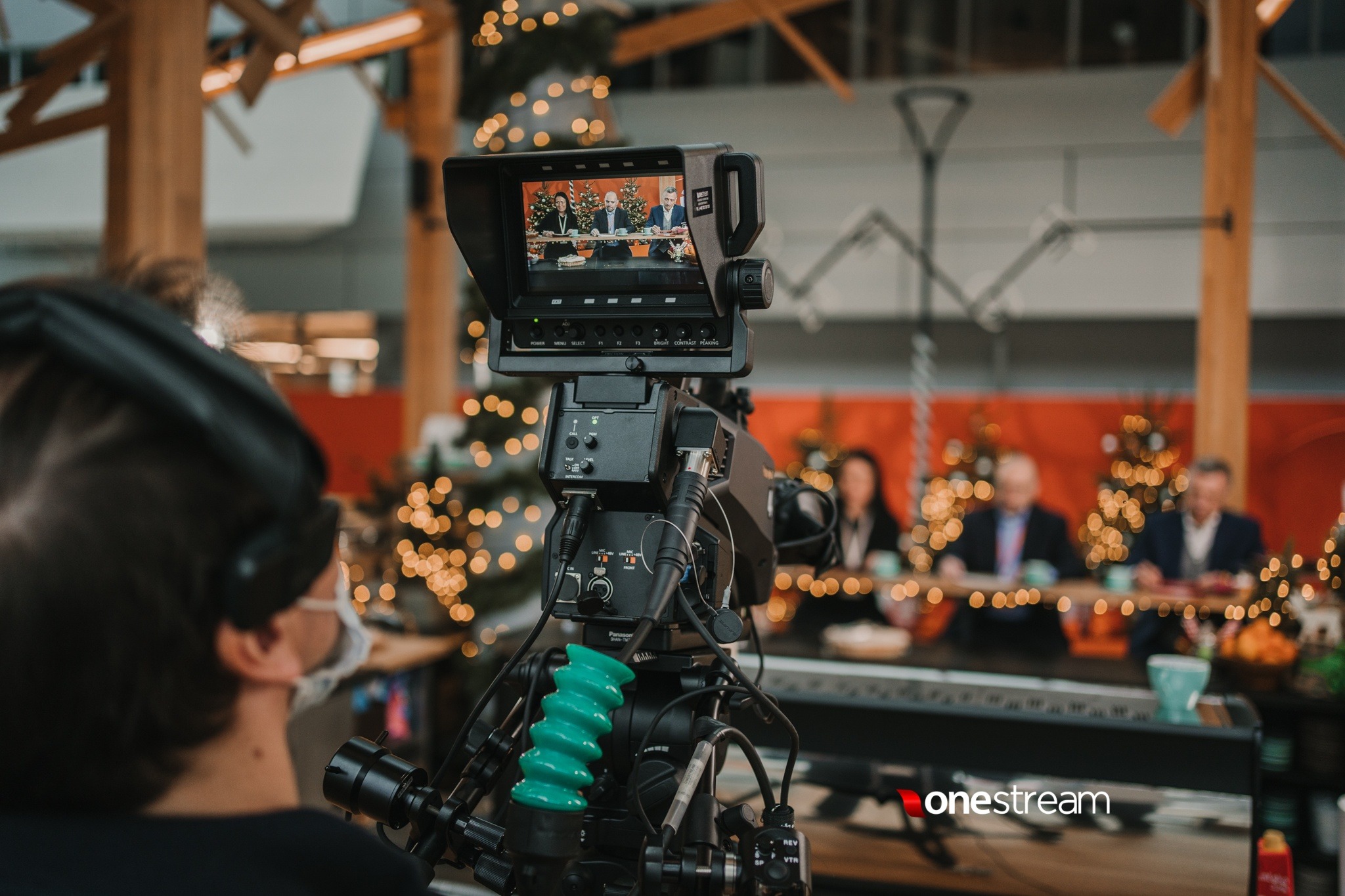 Kamera w studiu podczas nagrywania panelu dyskusyjnego z trzema osobami siedzącymi za stołem, w tle choinki z lampkami.