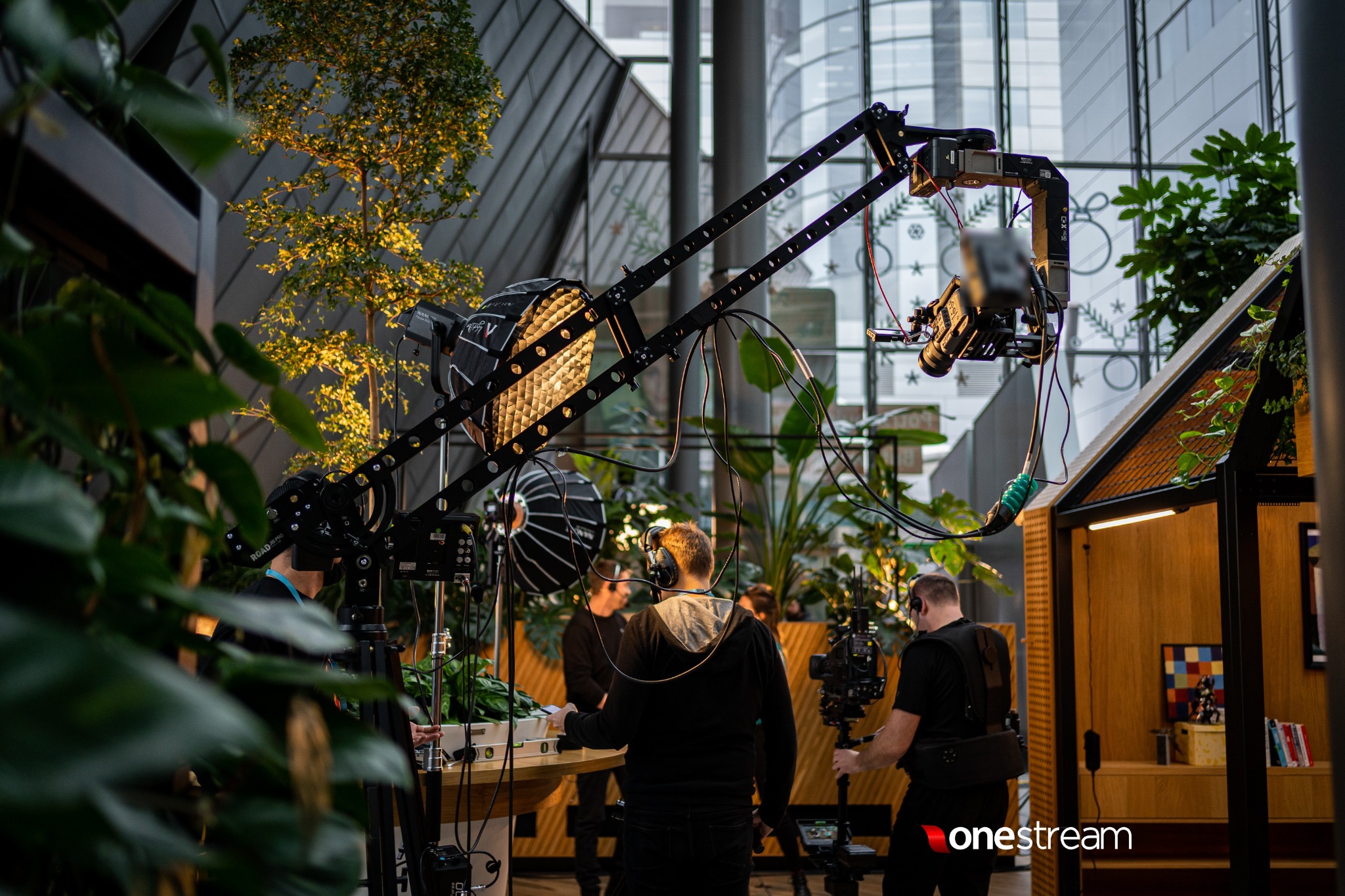 Ekipa filmowa podczas pracy w nowoczesnym wnętrzu z roślinami, widoczny wysięgnik kamery, oświetlenie i drewniana konstrukcja w tle.