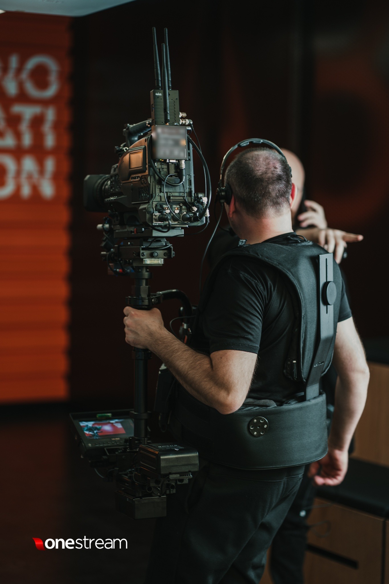 Operator kamery z zamontowanym rigiem i słuchawkami na głowie, widok z tyłu, w tle rozmyty napis na ścianie.