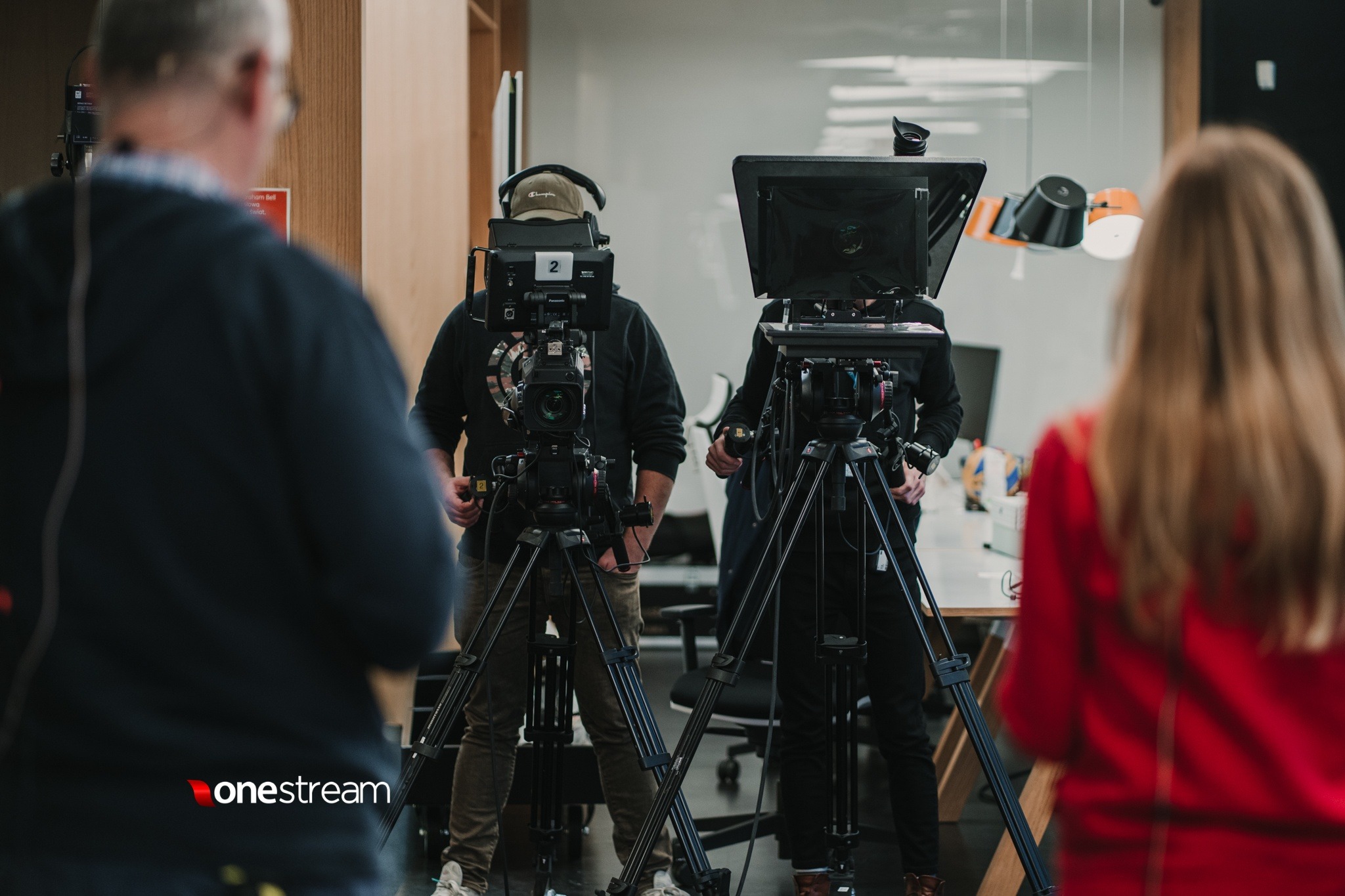 Dwaj kamerzyści z profesjonalnym sprzętem na statywach, widok zza pleców osoby w czerwonej bluzce, logo 'onestream' w lewym dolnym rogu.