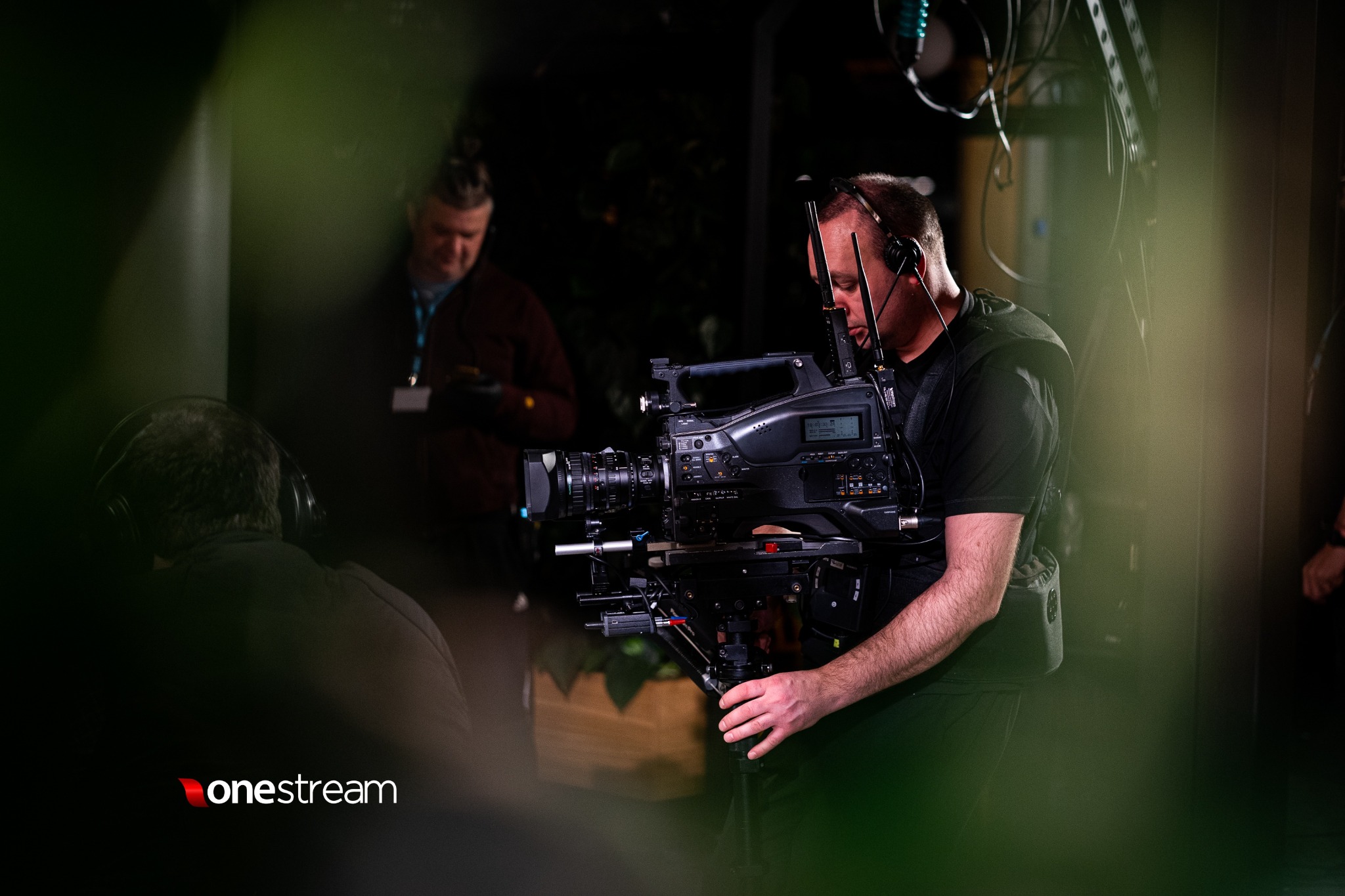 Kamerzysta w słuchawkach obsługuje profesjonalną kamerę w trakcie nagrania, widoczne okablowanie i elementy scenografii w tle.