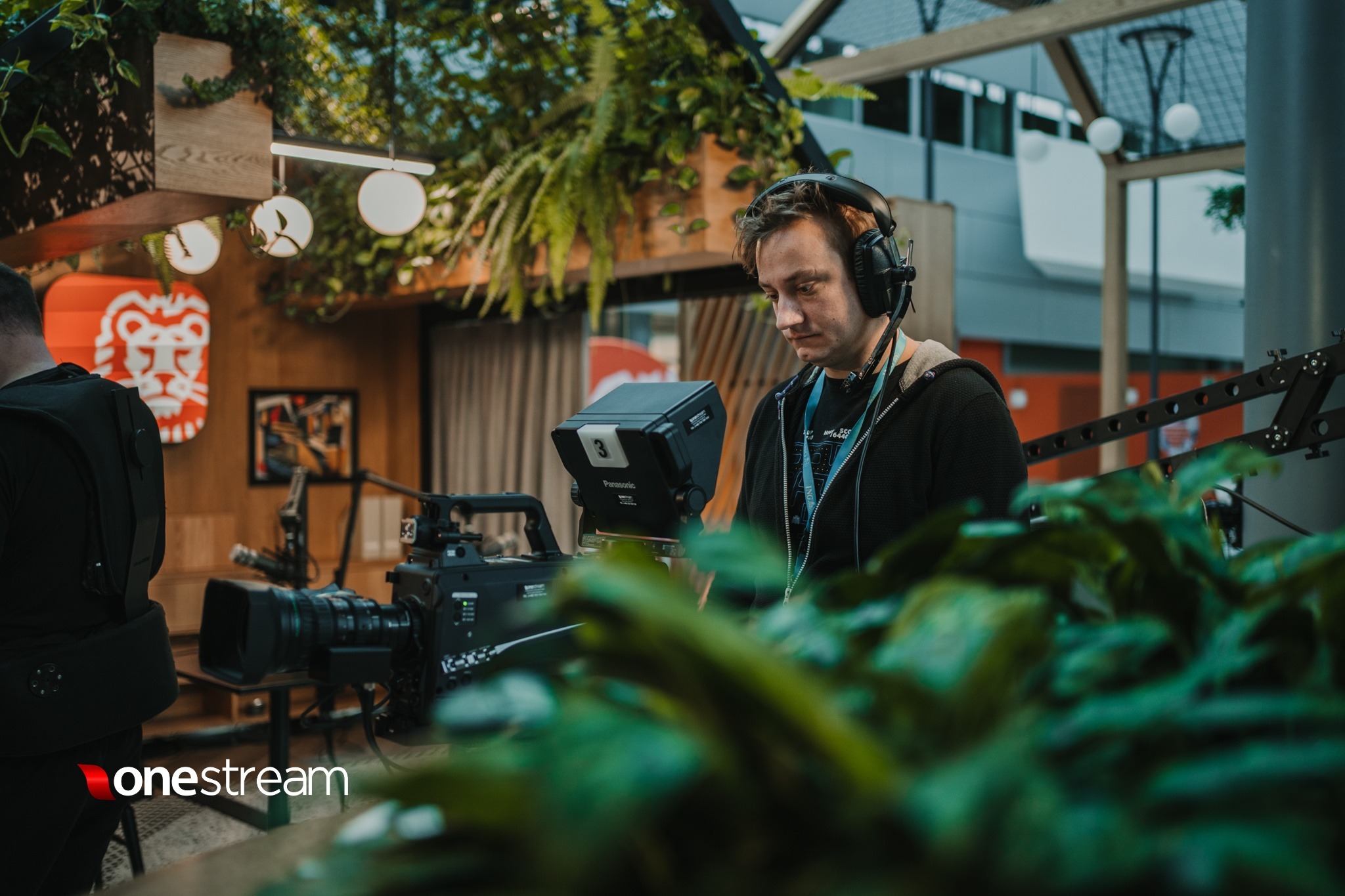 Operator filmowy w słuchawkach, obsługujący profesjonalną kamerę Panasonic, z widocznym logotypem firmy OneStream w tle, w otoczeniu roślinności i dekoracji wnętrz.