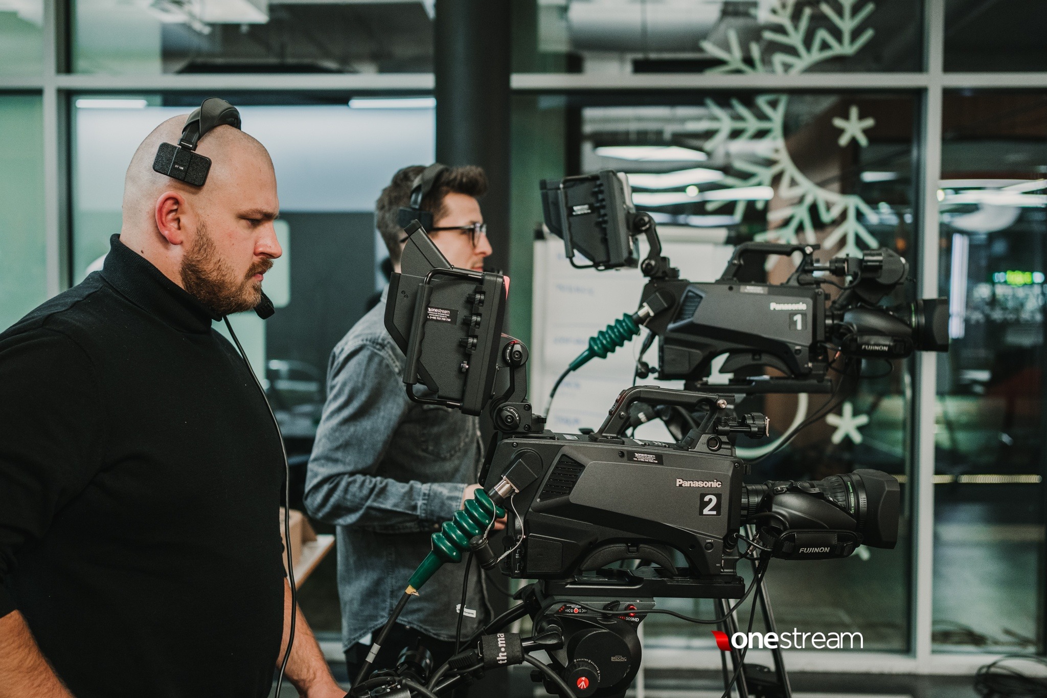 Dwóch mężczyzn w słuchawkach obsługuje profesjonalne kamery Panasonic na statywach, widoczne okno z dekoracją świąteczną w tle.