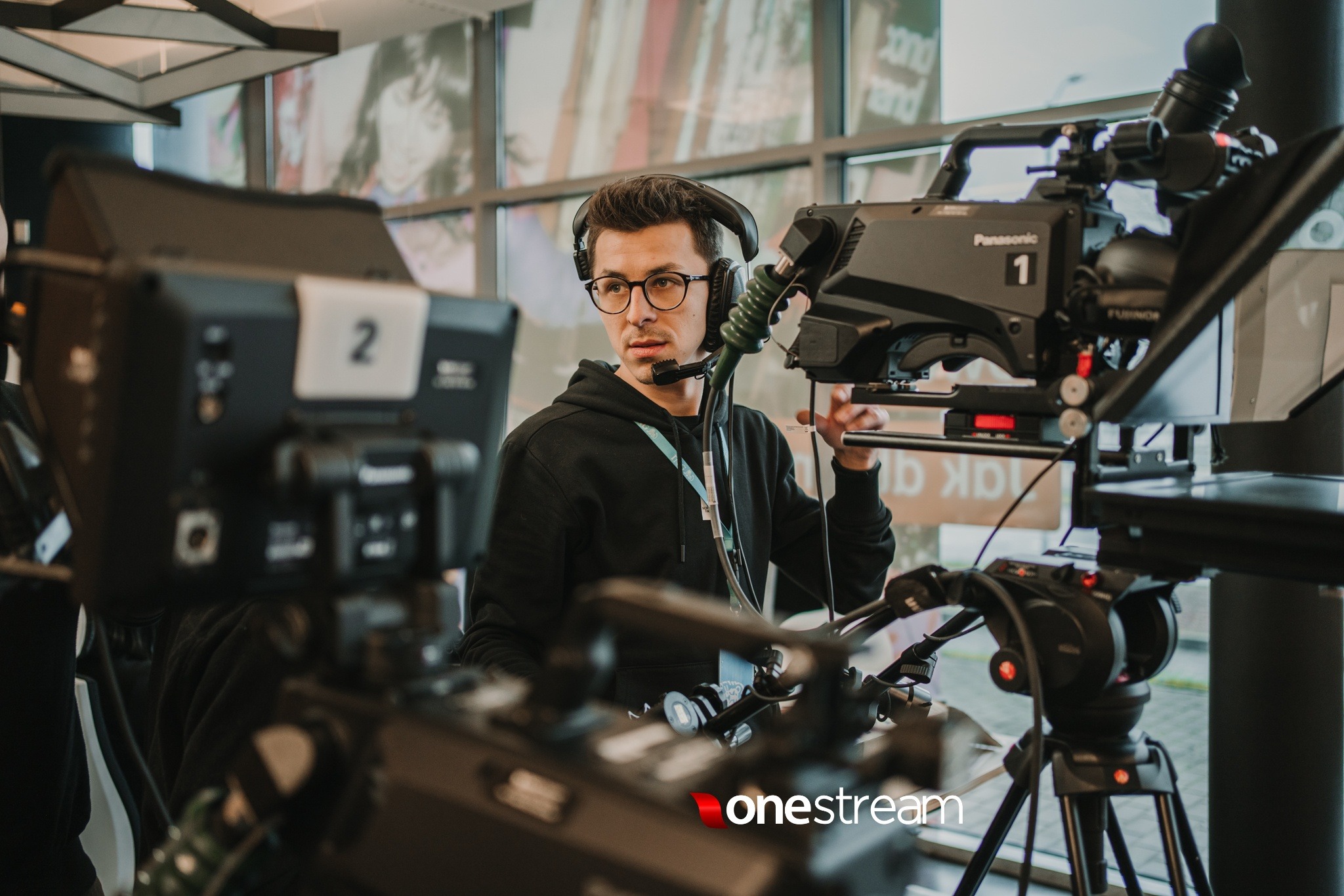 Kamerzysta w okularach i słuchawkach z mikrofonem, obsługujący profesjonalną kamerę Panasonic na statywie, w tle okno z plakatem.