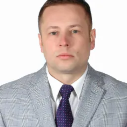 Portret mężczyzny w szarej kraciastej marynarce i granatowym krawacie w kropki na białym tle.