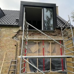 Magmax - Dom z cegły z zamontowanym oknem dachowym, widoczne rusztowanie i uszkodzona ściana zewnętrzna w trakcie naprawy.