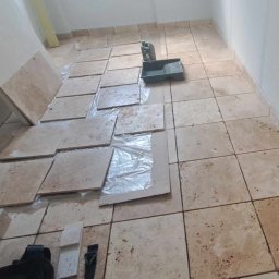 Magmax - Kładzenie beżowych, kwadratowych płytek podłogowych z widocznym pojemnikiem na farbę i folią ochronną na części płytek.